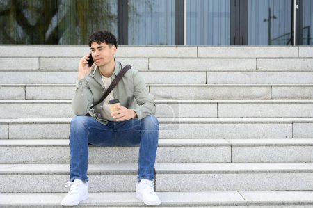 Téléchargez les photos : Attrayant confiant jeune homme d'affaires portant des vêtements décontractés, assis à l'extérieur parlant sur un téléphone mobile tout en buvant du café à emporter, scène urbaine de fond. - en image libre de droit