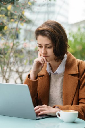 Foto de Foto vertical de una joven estudiante concentrada usando un portátil en una cafetería al aire libre - Imagen libre de derechos