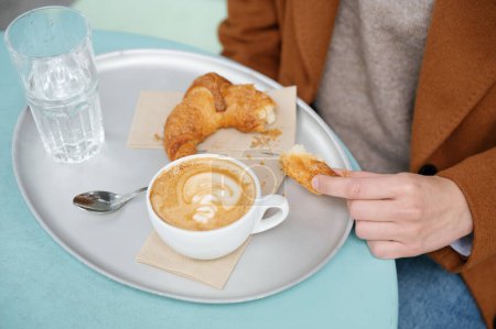 Téléchargez les photos : Gros plan d'une femme méconnaissable mangeant un croissant pendant le petit déjeuner assis dans une cafétéria extérieure - en image libre de droit