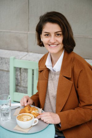 Photo for Vertical photo of a smiling woman having a delicious breakfast of croissant and coffee in a sidewalk cafeteria - Royalty Free Image