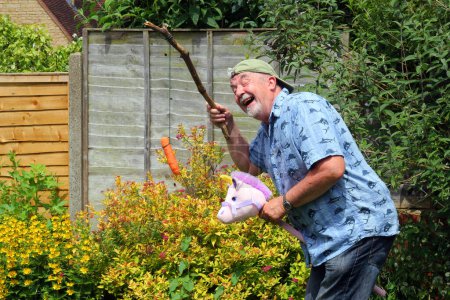 Photo for Elderly or senior man playing and having fun in his garden. Pretending to ride a horse with a carrot on a stick. Joking and enjoying himself. - Royalty Free Image