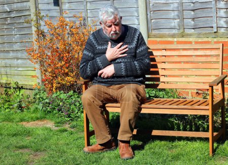 Photo for Elderly or senior man sitting on a bench with pains in his chest. Clutching his chest. possible heart attack. - Royalty Free Image