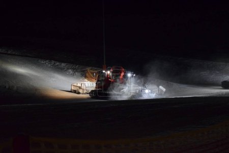 Téléchargez les photos : Un batteur de pistes travaillant au compactage de la neige dans une station de ski la nuit. - en image libre de droit