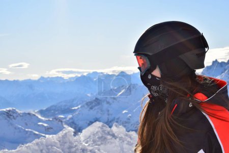 Female skier in winter sports gear with long hair, a black helmet, orange goggles and black face mask. Backdropped by snowcapped Italian mountain peaks of the Pennine alps on a clear, sunny day. 