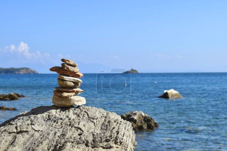 Balancierter Felsblock auf einem großen, groben Felsbrocken an einem Strand auf der griechischen Insel Skiathos. Im Hintergrund das helle, ruhige Ägäische Meer und ein klarer blauer Himmel.
