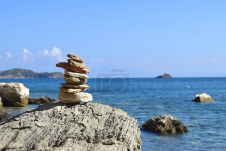 Balancierter Felsblock auf einem großen, groben Felsbrocken an einem Strand auf der griechischen Insel Skiathos. Im Hintergrund das helle, ruhige Ägäische Meer und ein klarer blauer Himmel.