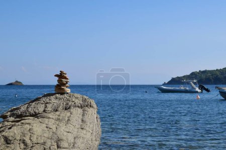 Piedra balanceada en una roca grande y áspera en una playa en la isla de Skiathos, Grecia. Trasladado por barcos en el tranquilo mar Egeo, con un cielo azul claro y brillante.