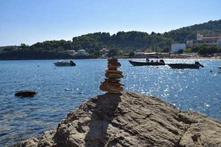 Balanced rock stack on a large, rough boulder on a beach on Skiathos island, Greece. Backdropped by boats on the calm aegean sea, with a bright, clear blue sky.