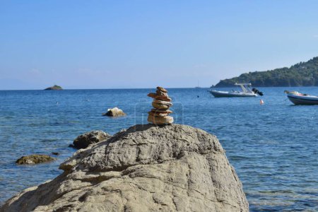 Balanced rock stack on a large, rough boulder on a beach on Skiathos island, Greece. Backdropped by boats on the calm aegean sea, with a bright, clear blue sky.
