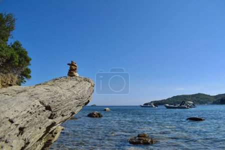 Balancierter Felsblock auf einem großen, groben Felsbrocken an einem Strand auf der Insel Skiathos, Griechenland. Im Hintergrund Boote auf der ruhigen Ägäis, mit einem hellen, klaren blauen Himmel.