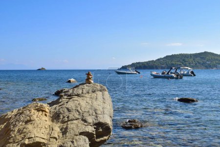 Balancierter Felsblock auf einem großen, groben Felsbrocken an einem Strand auf der Insel Skiathos, Griechenland. Im Hintergrund Boote auf der ruhigen Ägäis, mit einem hellen, klaren blauen Himmel.