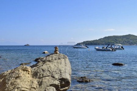 Foto de Piedra balanceada en una roca grande y áspera en una playa en la isla de Skiathos, Grecia. Trasladado por barcos en el tranquilo mar Egeo, con un cielo azul claro y brillante. - Imagen libre de derechos