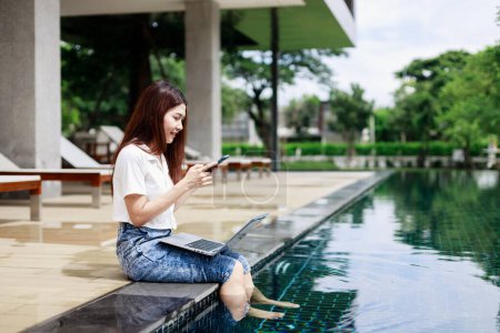 Téléchargez les photos : Une femme utilisant un smartphone pendant des vacances près de la piscine dans un hôtel de villégiature - en image libre de droit