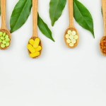 Top view of capsules and pills in wooden spoons with medicinal plant leaves on light background. Border composition with copy space