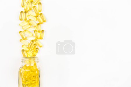 Yellow soft gel oil capsules spilled out of glass bottle on white background
