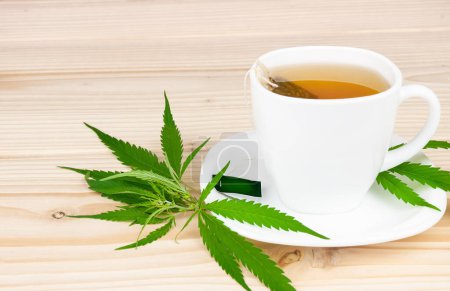 White cup with cannabis tea bag cannabis leaves on wooden table, copy space