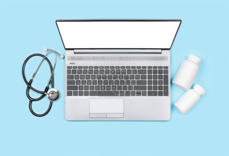 Top view of laptop with blank display, pill bottles and stethoscope on blue background. Telemedicine concept