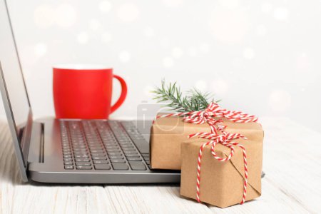 Foto de Cajas de regalo de Navidad y taza cerca de la computadora portátil en la mesa blanca. Lugar de trabajo con luces desenfocadas y espacio de copia. Compras en línea o concepto de saludo - Imagen libre de derechos