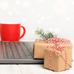 Christmas gift boxes and cup near the laptop on white table. Working place with defocused lights and copy space. Online shopping or greeting concept