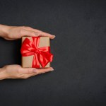 Female hands holding craft gift box with red ribbon on black background. Top view with copy space