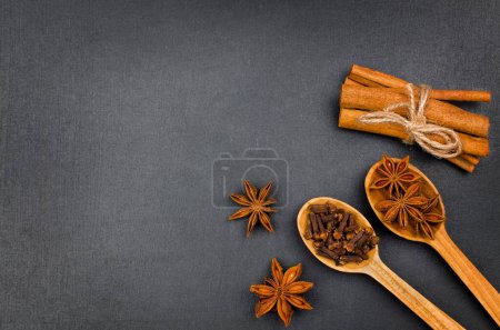 Cinnamon sticks, star anise and cloves in wooden spoons on black background. Top view with copy space, flat lay