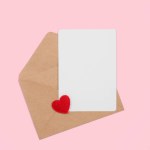 Open craft envelope with empty postcard and heart on pink pastel background, top view with copy space