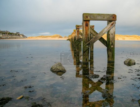 Téléchargez les photos : Un vieux squelette de pont en bois, vieux pont historique en bois soutient à travers tranquille, encore rivière à la plage de sable en été - en image libre de droit