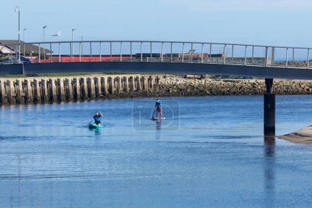 Foto de Pareja relajada disfrutando de un tranquilo paddle boarding en aguas tranquilas con un puente y una hermosa reflexión en un día soleado de vacaciones cerca de la playa - Imagen libre de derechos