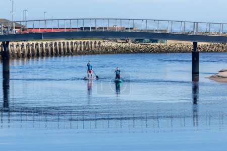 Photo for Relaxed couple enjoying tranquil paddle boarding on still water with a bridge and lovely reflection on a sunny day on holiday near the beach - Royalty Free Image