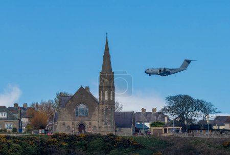 An Atlas RAF carrier aeroplane, aircraft low flying over the town preparing to land with a church spire in a blue sky