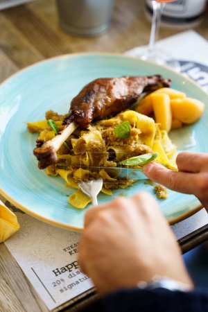 A person eating roasted goose leg with cabbage papardelle pasta and quince compote