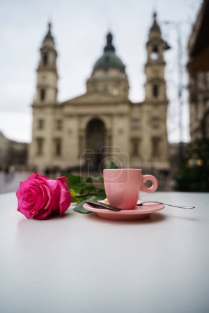 Una rosa rosada y una taza de café en una mesa frente a la basílica