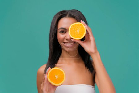 Photo for Natural spa. Beautiful young African American shirtless woman holding pieces of orange in front of her eyes while standing isolated on turquoise background - Royalty Free Image