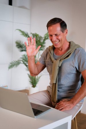 Photo for Mature man using laptop, making video call, sitting at table in kitchen, senior teacher mentor wearing glasses engaged online conference, recording webinar, teaching - Royalty Free Image