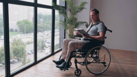 Téléchargez les photos : Homme adulte en fauteuil roulant lisant un livre près de la fenêtre de la maison - en image libre de droit