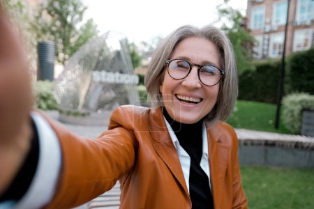 Téléchargez les photos : Moyen âge femme aux cheveux gris souriant heureux faire selfie par la caméra à la ville - en image libre de droit