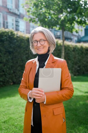 Photo for Confident mature business woman with a laptop in her hands on the background of a city park, wearing stylish eyeglasses standing. Portrait of smiling manager looking at the camera. - Royalty Free Image