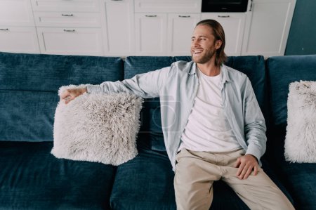 Foto de Un hombre guapo se sienta en casa en el sofá y sonríe. Una rubia con barba se enfría en un cómodo sofá - Imagen libre de derechos