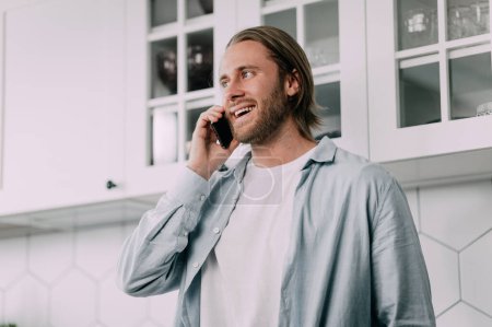 Foto de Hombre rubio hablando por teléfono en la cocina en casa y sonriendo. - Imagen libre de derechos