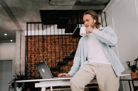 Téléchargez les photos : L'homme boit du café sur le lieu de travail Dans un bureau élégant ou un espace de coworking, un employé se refroidit avec une tasse de thé. - en image libre de droit