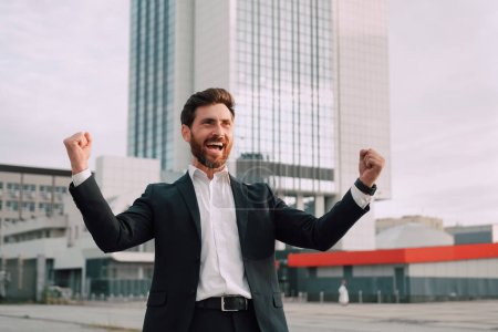 Photo for Happy millennial bearded businessman in suit rejoices in successful deal or win, rises hands up near modern office building, outdoor, emotions of victory. - Royalty Free Image