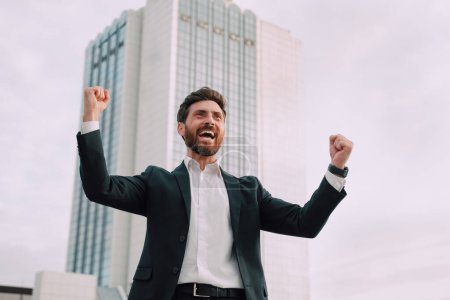 Happy satisfied excited european millennial bearded businessman in suit rejoices in successful deal or win, rises hands up near modern office building, outdoor. Manager, emotions of victory, gestures.