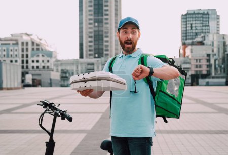 Téléchargez les photos : Un livreur de nourriture avec un scooter et pizza regarde sa montre et se rend compte qu'il est en retard, choqué. - en image libre de droit