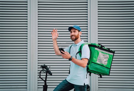 Photo for Friendly courier pizza delivery man with thermal backpack and phone on a E-Scooter. Fast transport express home delivery. Online order. Delivery man in cap. - Royalty Free Image