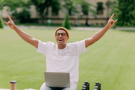 Téléchargez les photos : Élégant homme afro-américain branché se réjouit du succès avec un ordinateur portable à l'extérieur sur un banc. - en image libre de droit