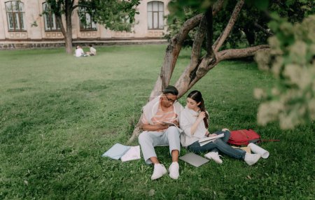 Téléchargez les photos : Quelques étudiants internationaux travaillant sur des devoirs ensemble assis sur la pelouse près de l'arbre. - en image libre de droit