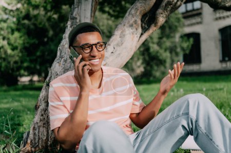 Téléchargez les photos : Élégant jeune homme afro-américain assis sur une pelouse sous un arbre et parlant au téléphone et souriant joyeusement. - en image libre de droit
