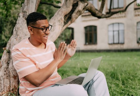Téléchargez les photos : Jeune homme multiethnique élégant assis sur la pelouse ayant une vidéoconférence avec un ordinateur portable - en image libre de droit