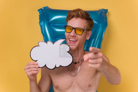 Photo for Man with inflatable beach mattress showing speech bubble and approving gesture. Summer vacation concept on yellow background. - Royalty Free Image