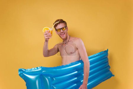 Téléchargez les photos : Heureux homme torse nu drôle tenant verre à cocktail avec du jus d'orange frais et matelas gonflable sur fond jaune - en image libre de droit